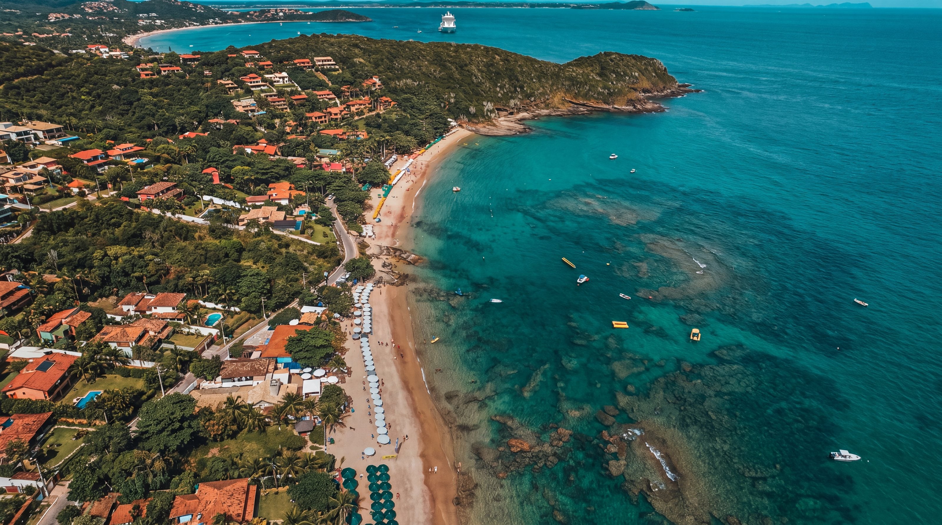 Vista aérea de uma praia de Búzios, com mar azul-turquesa, guarda-sóis e encosta verde