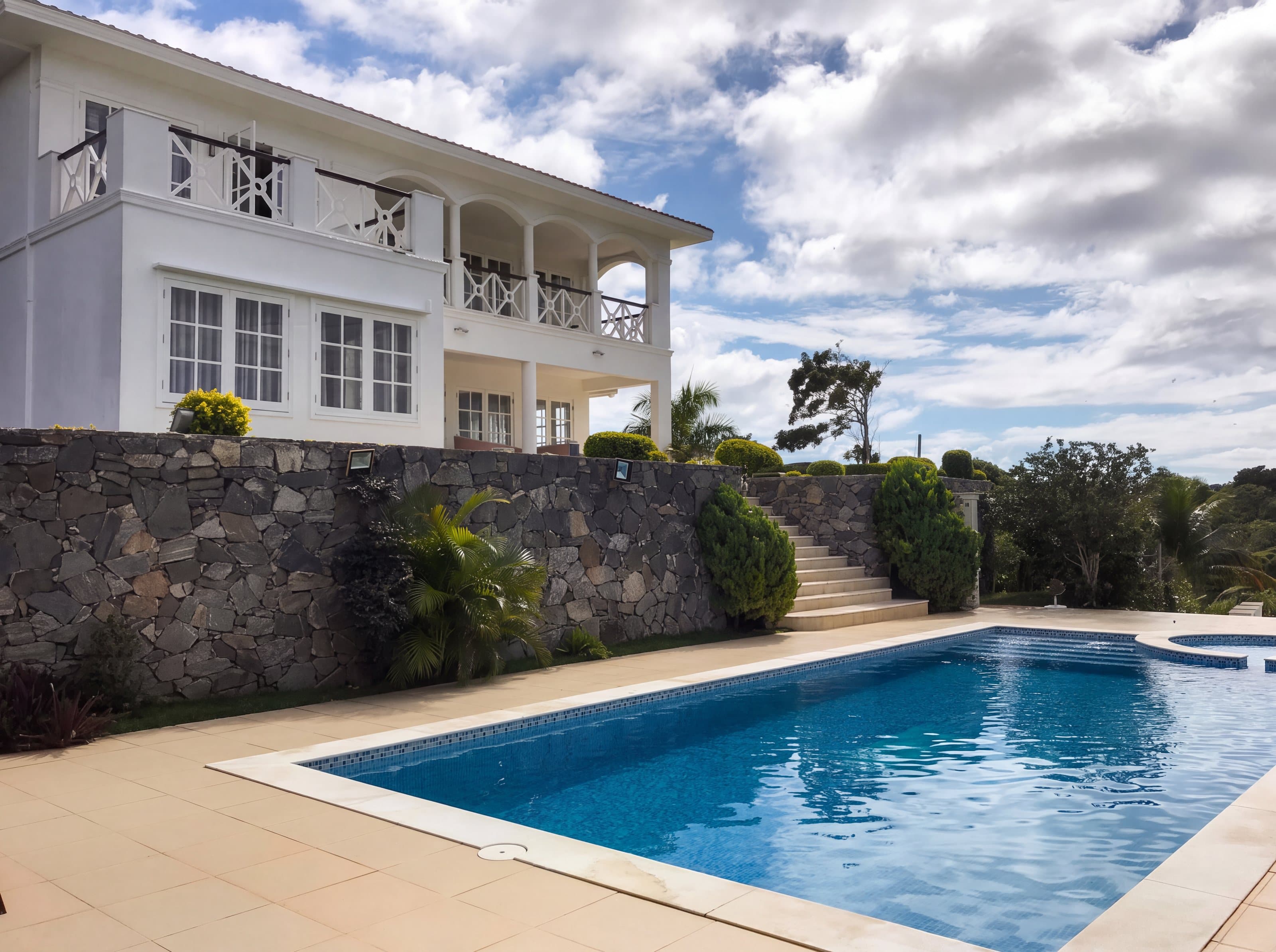 Casa branca com varanda em arcos sobre muro de pedra, piscina azul e céu com nuvens